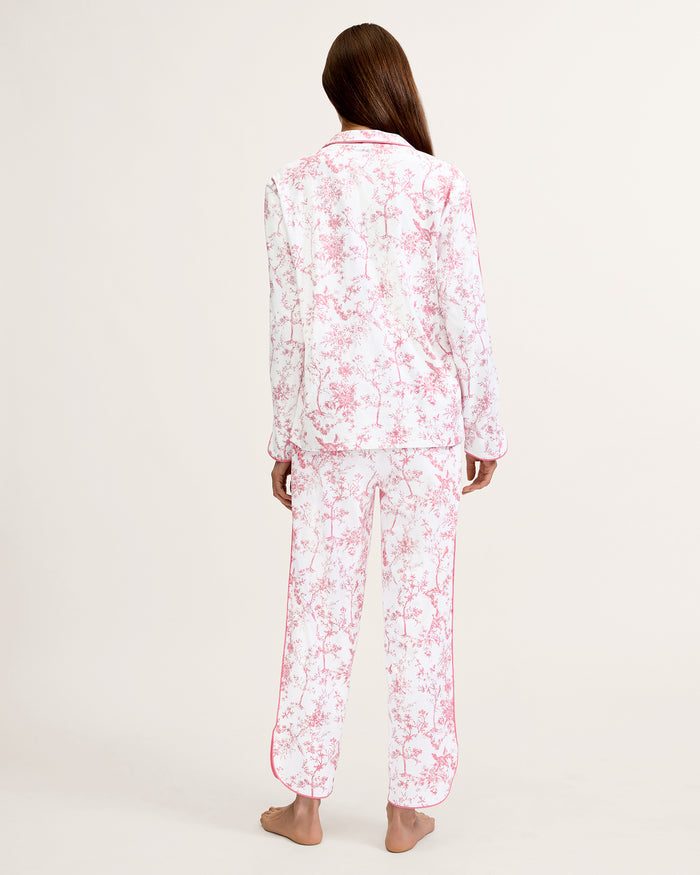 A woman stands barefoot on a plain background, wearing The Margaux Pima Pajama Set in Pink Timeless Toile. She has straight brown hair and lightly touches her chin with one hand.