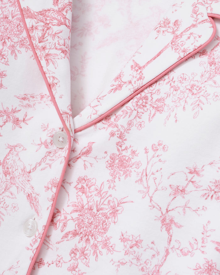 A woman stands barefoot on a plain background, wearing The Margaux Pima Pajama Set in Pink Timeless Toile. She has straight brown hair and lightly touches her chin with one hand.
