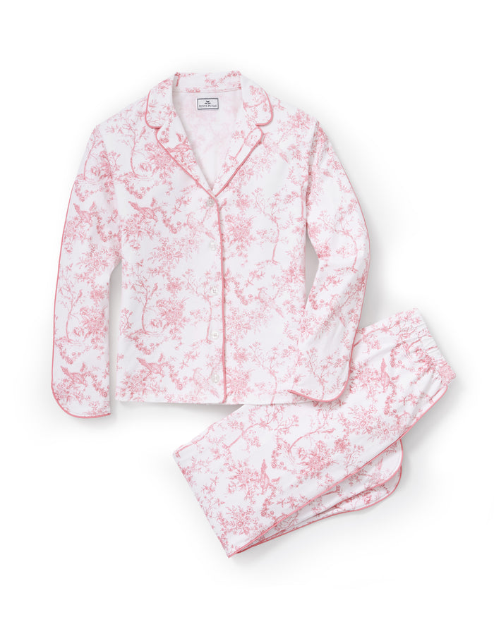 A woman stands barefoot on a plain background, wearing The Margaux Pima Pajama Set in Pink Timeless Toile. She has straight brown hair and lightly touches her chin with one hand.