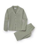 A woman with long dark hair wears The Margaux Pima Pajama Set in Sage, featuring a button-up long-sleeve top with white piping and matching pants. She stands barefoot against a plain, light background.