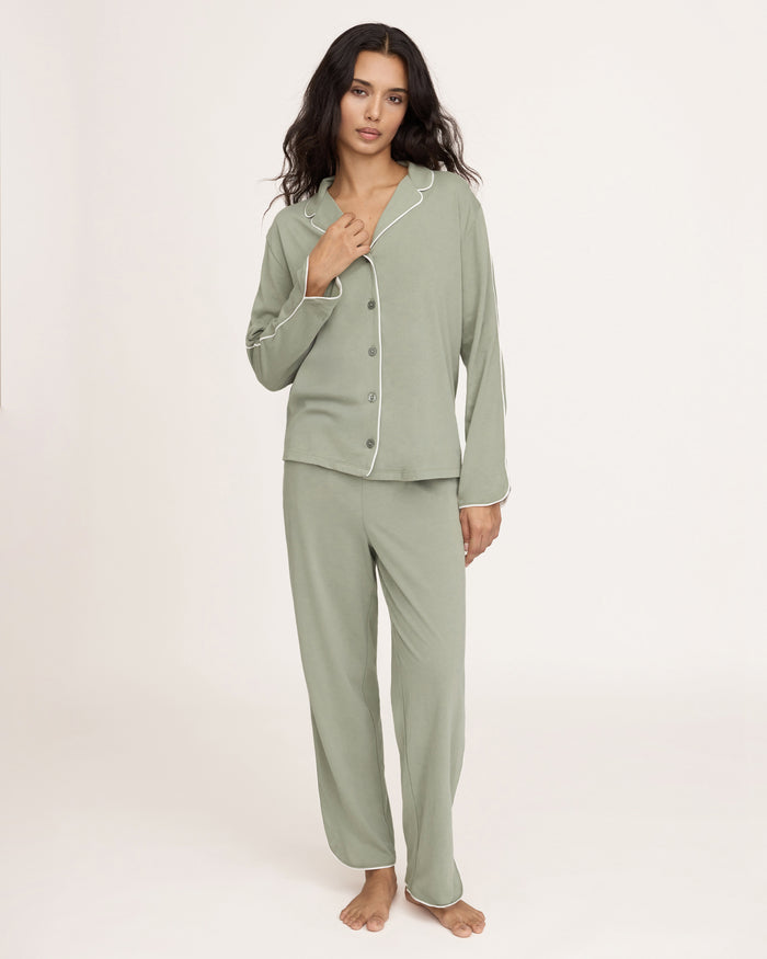 A woman with long dark hair wears The Margaux Pima Pajama Set in Sage, featuring a button-up long-sleeve top with white piping and matching pants. She stands barefoot against a plain, light background.