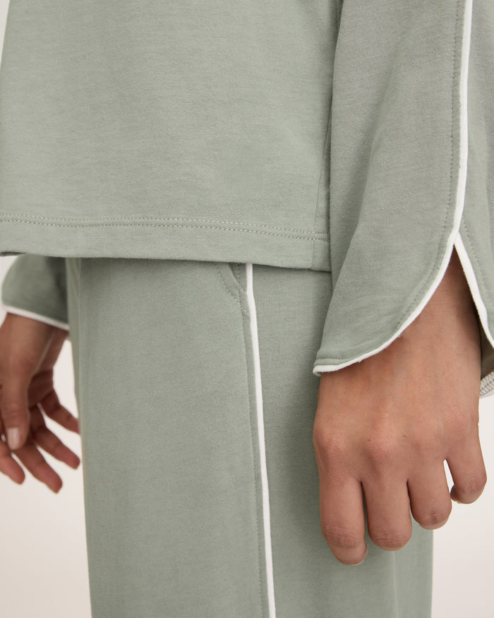 A woman with long dark hair wears The Margaux Pima Pajama Set in Sage, featuring a button-up long-sleeve top with white piping and matching pants. She stands barefoot against a plain, light background.