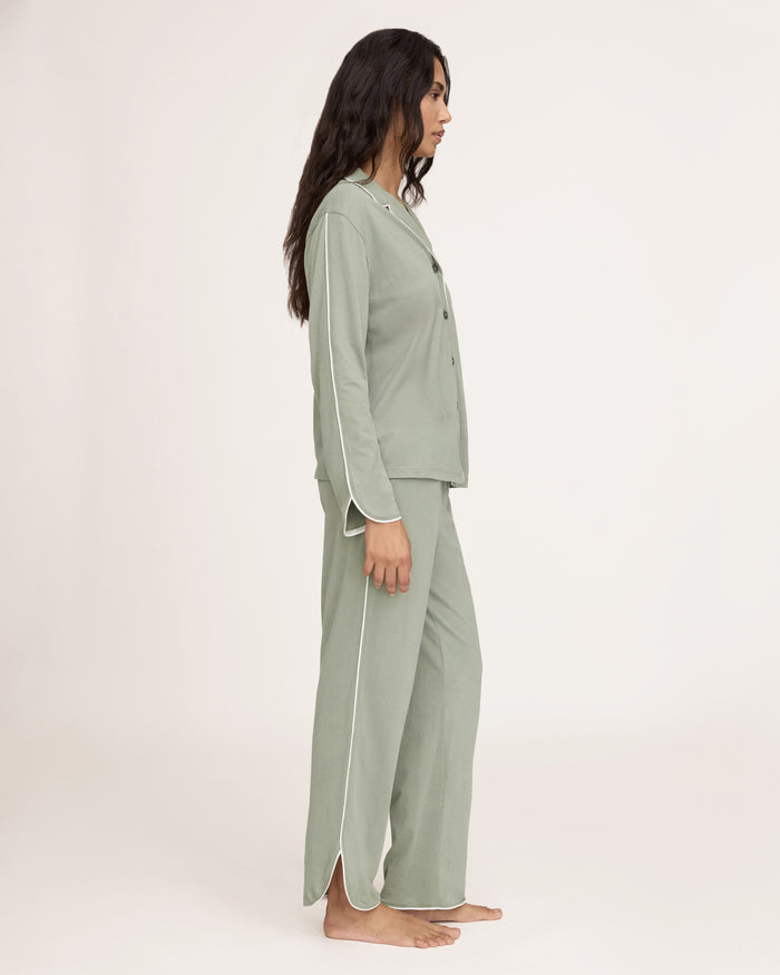 A woman with long dark hair wears The Margaux Pima Pajama Set in Sage, featuring a button-up long-sleeve top with white piping and matching pants. She stands barefoot against a plain, light background.