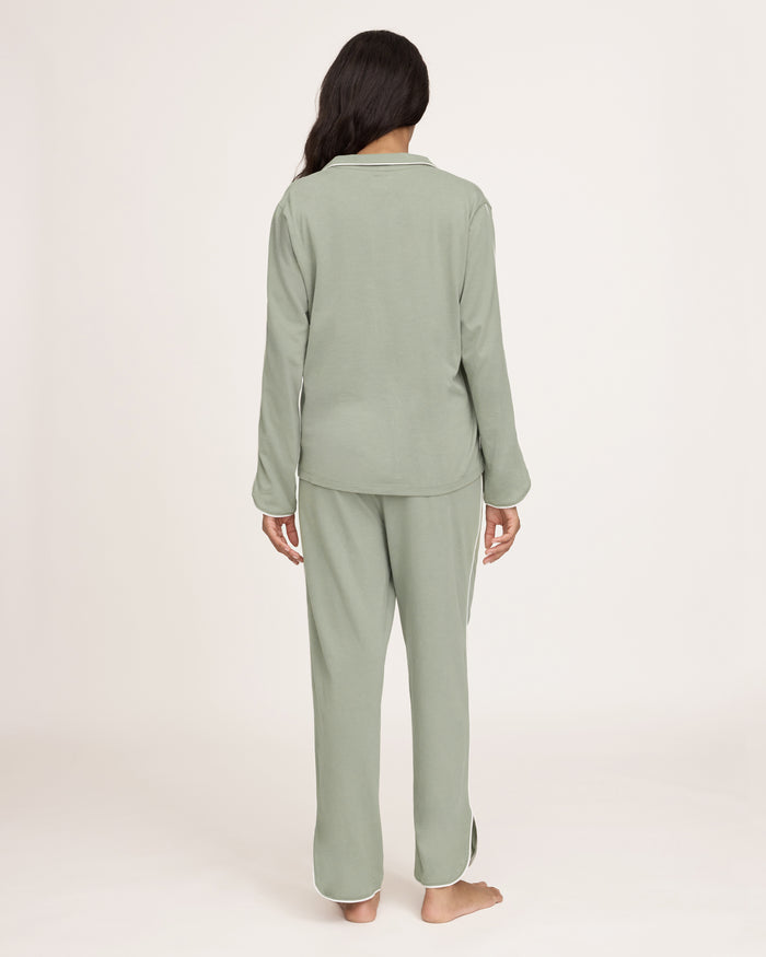 A woman with long dark hair wears The Margaux Pima Pajama Set in Sage, featuring a button-up long-sleeve top with white piping and matching pants. She stands barefoot against a plain, light background.