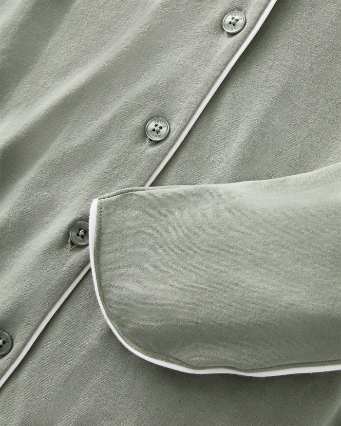 A woman with long dark hair wears The Margaux Pima Pajama Set in Sage, featuring a button-up long-sleeve top with white piping and matching pants. She stands barefoot against a plain, light background.