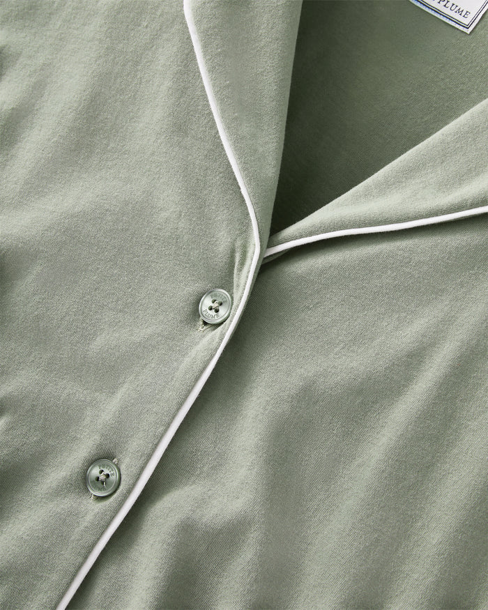 A woman with long dark hair wears The Margaux Pima Pajama Set in Sage, featuring a button-up long-sleeve top with white piping and matching pants. She stands barefoot against a plain, light background.