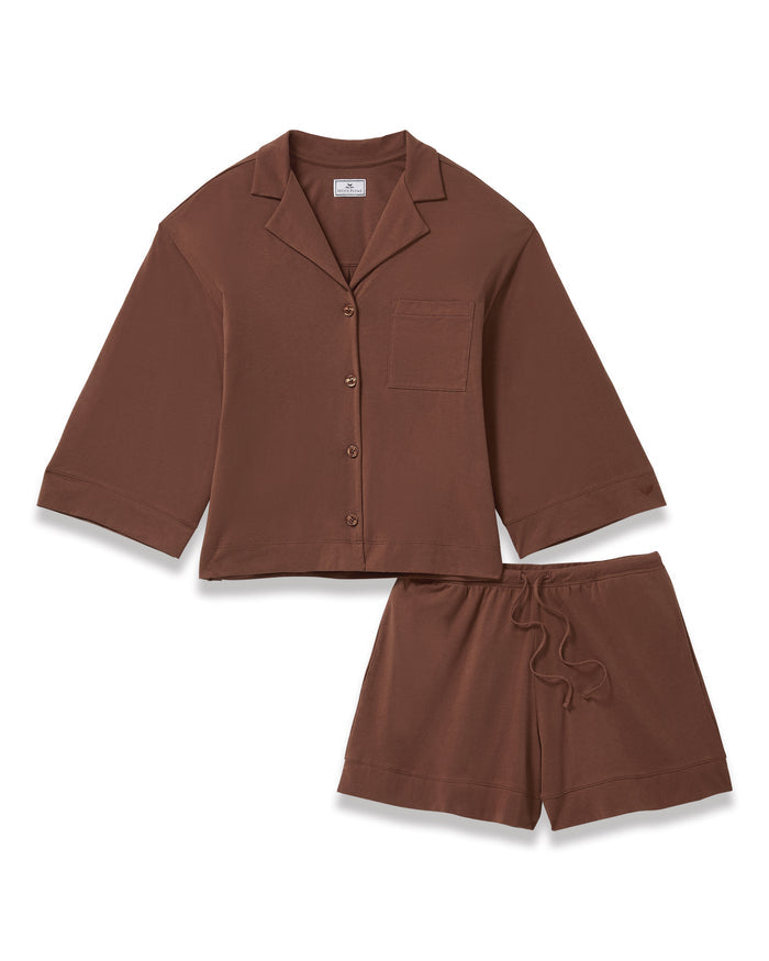A woman stands barefoot against a white background, wearing The Olivia Lightweight Pima Short Set in Chocolate Brown—featuring a loose-fitting, long-sleeved button-up top with a pocket and matching shorts.