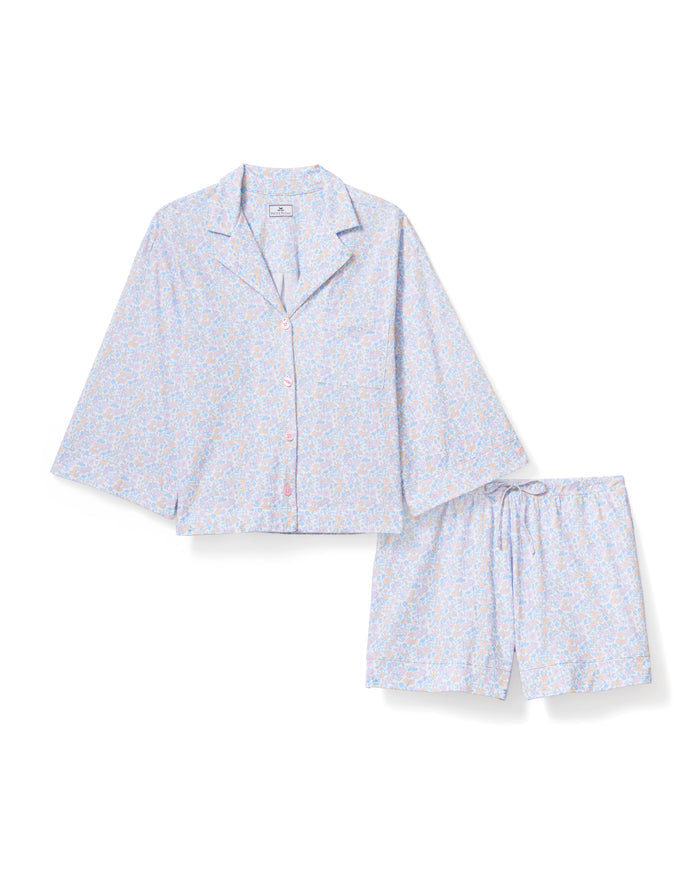 A woman with long, wavy hair wears The Olivia Lightweight Pima Short Set in Fleur Blanche, featuring a loose button-up top with wide sleeves and matching shorts adorned with a subtle floral pattern. She stands against a plain, light background.