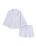 A woman with long, wavy hair wears The Olivia Lightweight Pima Short Set in Fleur Blanche, featuring a loose button-up top with wide sleeves and matching shorts adorned with a subtle floral pattern. She stands against a plain, light background.
