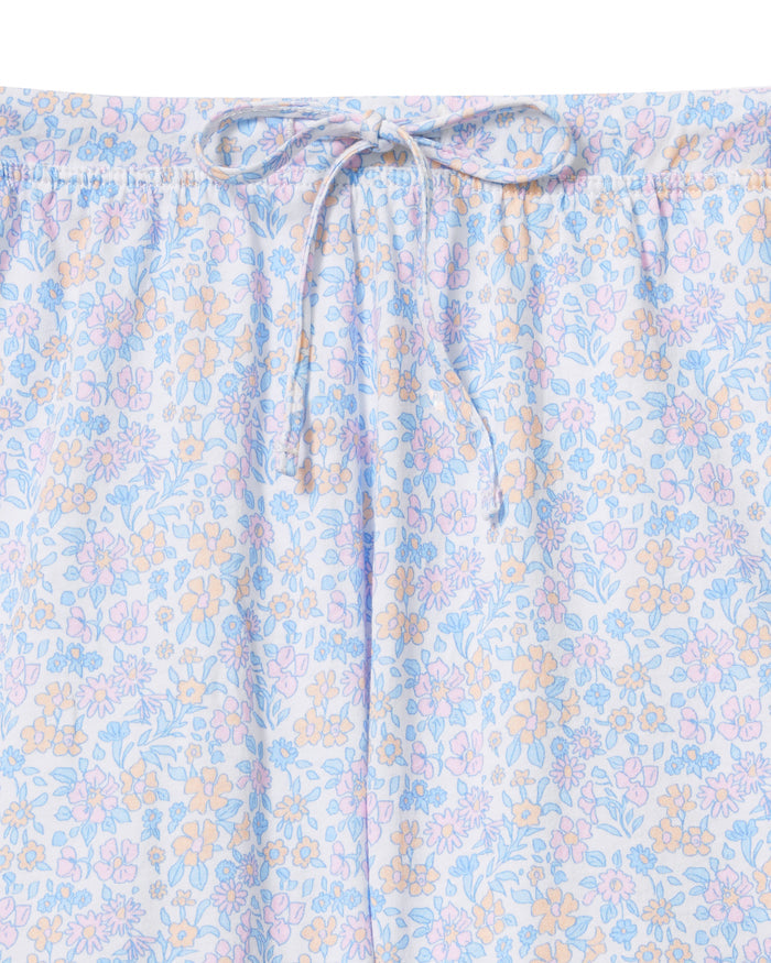 A woman with long, wavy hair wears The Olivia Lightweight Pima Short Set in Fleur Blanche, featuring a loose button-up top with wide sleeves and matching shorts adorned with a subtle floral pattern. She stands against a plain, light background.