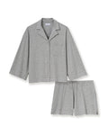 A woman with long, wavy hair and a neutral expression stands barefoot against a plain background, wearing The Olivia Lightweight Pima Short Set in Heather Grey—featuring a loose-fitting, long-sleeve top and matching shorts.
