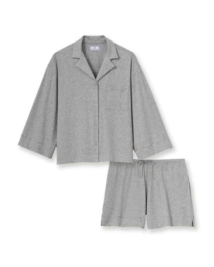 A woman with long, wavy hair and a neutral expression stands barefoot against a plain background, wearing The Olivia Lightweight Pima Short Set in Heather Grey—featuring a loose-fitting, long-sleeve top and matching shorts.