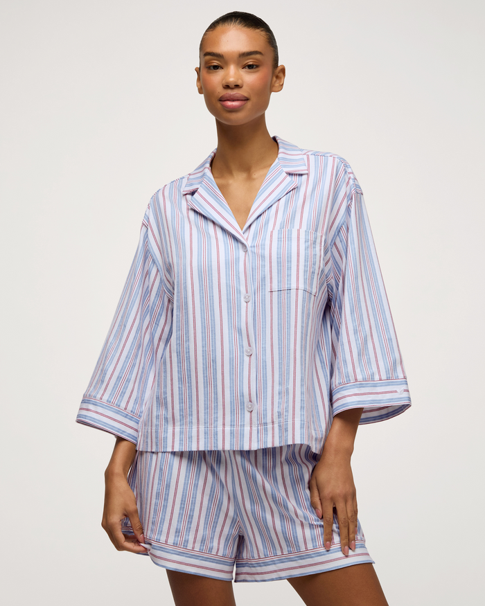 A woman stands against a plain light background, wearing The Olivia Lightweight Pima Short Set in Vintage French Stripes—a matching pajama set with blue, white, and red vertical stripes, featuring a button-up top with three-quarter sleeves and coordinating shorts.