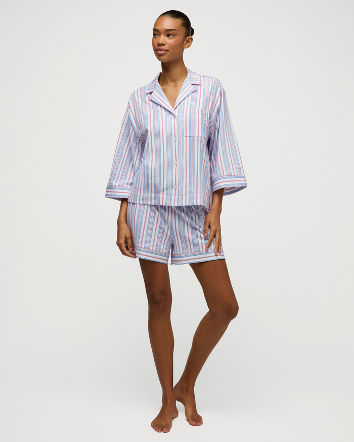 Standing barefoot against a plain white background, a person wears The Olivia Lightweight Pima Short Set in Vintage French Stripes—a matching set featuring loose, striped pajamas with a button-up top and shorts in blue, pink, and white tones.