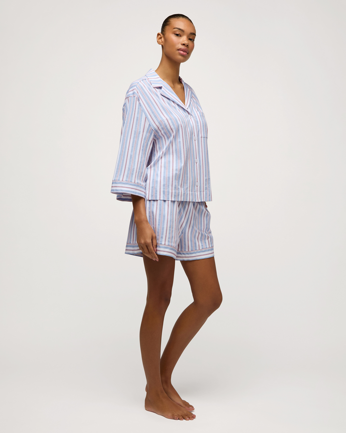 A person stands barefoot against a plain light background, wearing The Olivia Lightweight Pima Short Set in Vintage French Stripes—featuring a blue and white striped pajama set with long sleeves and shorts.
