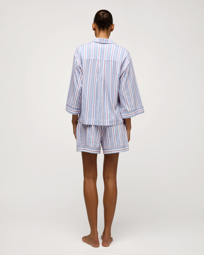 A person stands barefoot facing away, wearing The Olivia Lightweight Pima Short Set in Vintage French Stripes—a matching pajama set featuring blue, white, and red vertical stripes with a long-sleeve top and shorts. The background is plain and light.