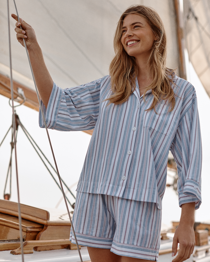 A smiling woman on a sailboat wears The Olivia Lightweight Pima Short Set in Vintage French Stripes, holding a rope with white sails in the background.