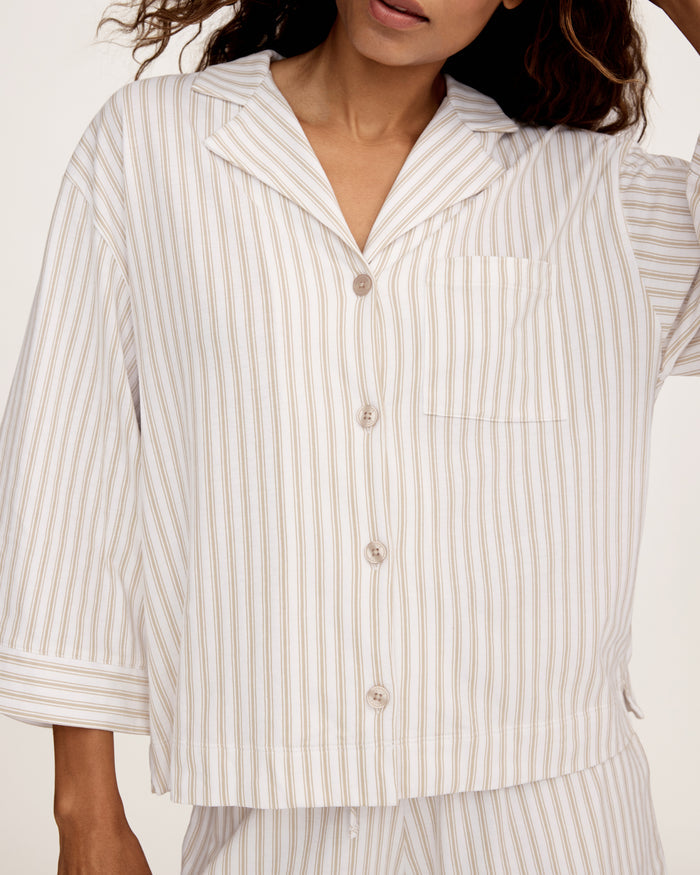 The Olivia Pima Short Set in Camel Ticking features a two-piece pajama set with beige and white vertical stripes, including a button-up, collared long-sleeve shirt with a breast pocket, and matching relaxed-fit drawstring shorts.