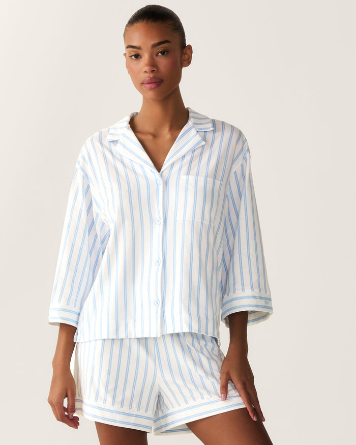 A woman wearing The Olivia Pima Short Set in Periwinkle Stripe—a matching pajama set with blue and white vertical stripes, featuring a button-up collared top with three-quarter sleeves and relaxed-fit shorts.