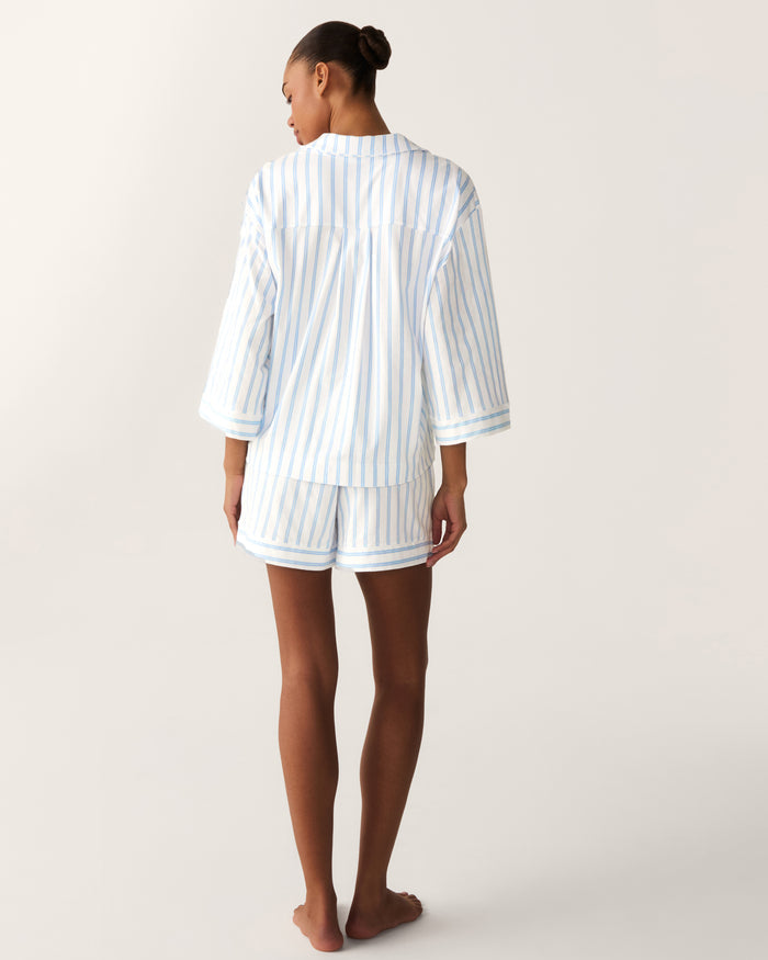 A person stands barefoot facing away, wearing The Olivia Pima Short Set in Periwinkle Stripe—a matching pajama set with blue and white vertical stripes, featuring a loose long-sleeve top and shorts—set against a plain background.