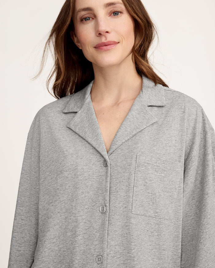 A woman stands barefoot against a plain background, smiling gently with one hand by her side, wearing The Olivia Wide Leg Lightweight Pima Pajama Set in Heather Grey—loose-fitting pants and a matching long-sleeve button-up top.