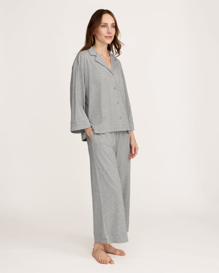 A woman stands barefoot against a plain background, smiling gently with one hand by her side, wearing The Olivia Wide Leg Lightweight Pima Pajama Set in Heather Grey—loose-fitting pants and a matching long-sleeve button-up top.