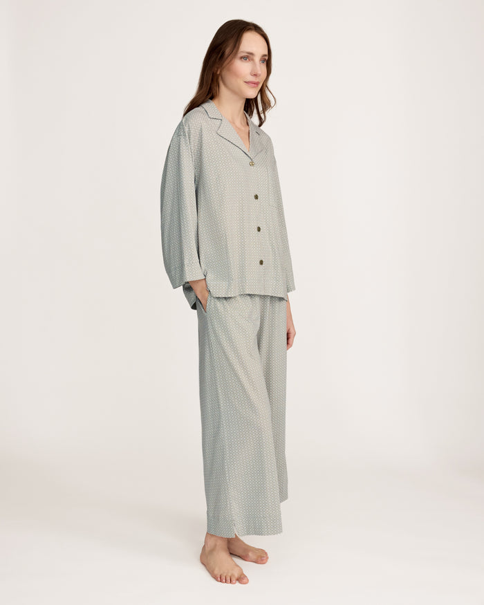 A woman stands barefoot against a plain white background, wearing The Olivia Wide Leg Lightweight Pima Pajama Set in Sage Geo—a matching loose-fitting ensemble with long sleeves and wide-leg pants in a subtle light pattern.