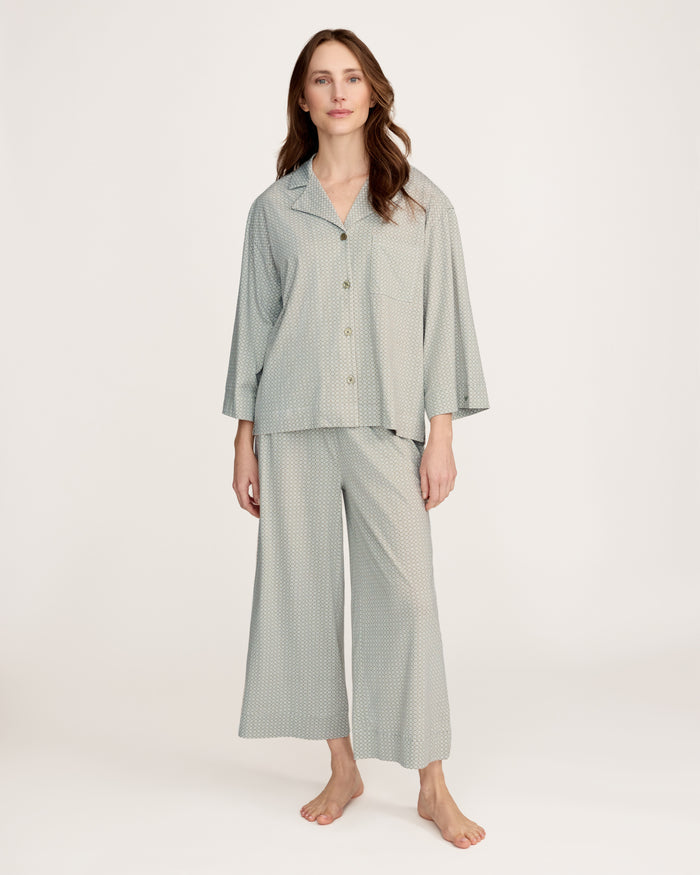 A woman stands barefoot against a plain white background, wearing The Olivia Wide Leg Lightweight Pima Pajama Set in Sage Geo—a matching loose-fitting ensemble with long sleeves and wide-leg pants in a subtle light pattern.
