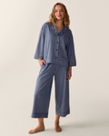 A person stands barefoot against a plain light background, wearing The Olivia Wide Leg Pima Pajama Set in Admiral Ticking, which features loose-fitting blue and white vertical stripes, a long-sleeved button-up top, and wide-leg pants.