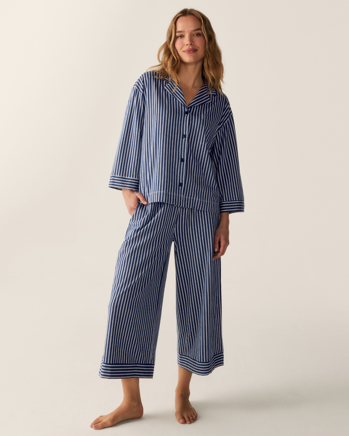A person stands barefoot against a plain light background, wearing The Olivia Wide Leg Pima Pajama Set in Admiral Ticking, which features loose-fitting blue and white vertical stripes, a long-sleeved button-up top, and wide-leg pants.