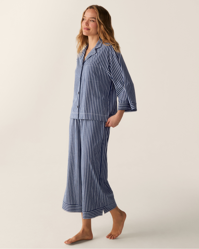 A woman stands barefoot against a plain light background, wearing The Olivia Wide Leg Pima Pajama Set in Admiral Ticking, featuring blue and white vertical stripes, long sleeves, and wide-leg pants.