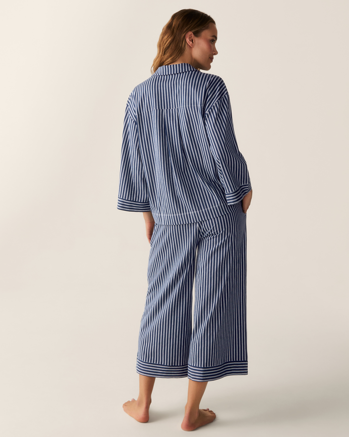 Wearing The Olivia Wide Leg Pima Pajama Set in Admiral Ticking, a woman stands barefoot with her back to the camera. The loose-fitting pajamas feature vertically striped blue and white fabric, cropped wide-leg pants, and long sleeves, all set against a plain light background.