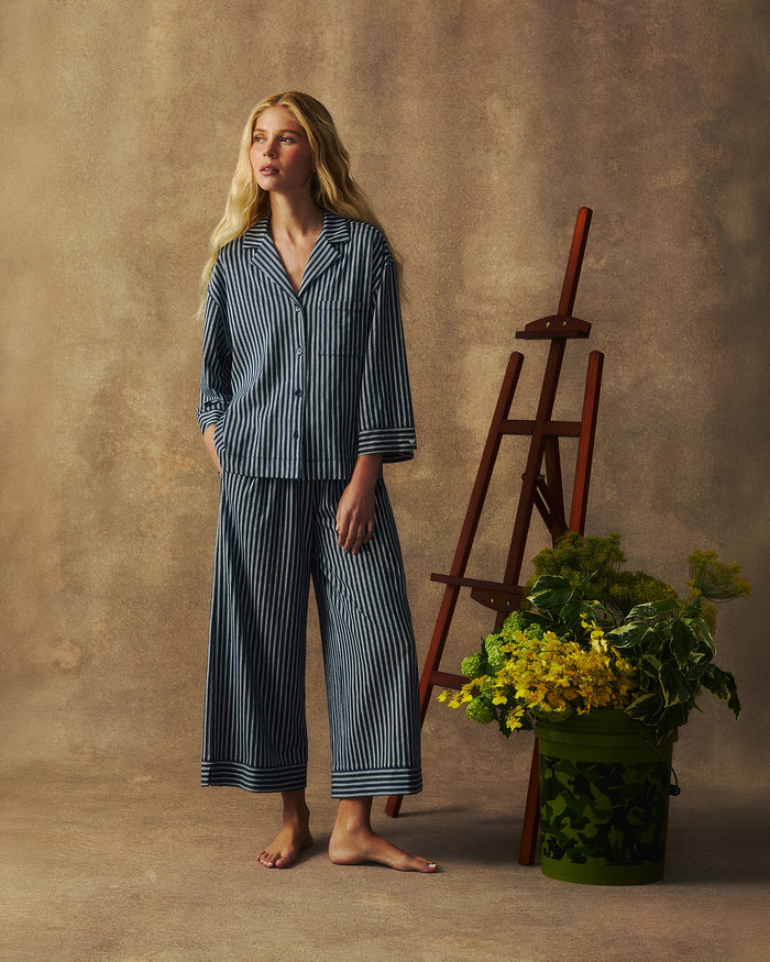 Wearing The Olivia Wide Leg Pima Pajama Set in Admiral Ticking, a barefoot woman stands by a wooden easel and a green basket filled with yellow flowers, set against a neutral backdrop.