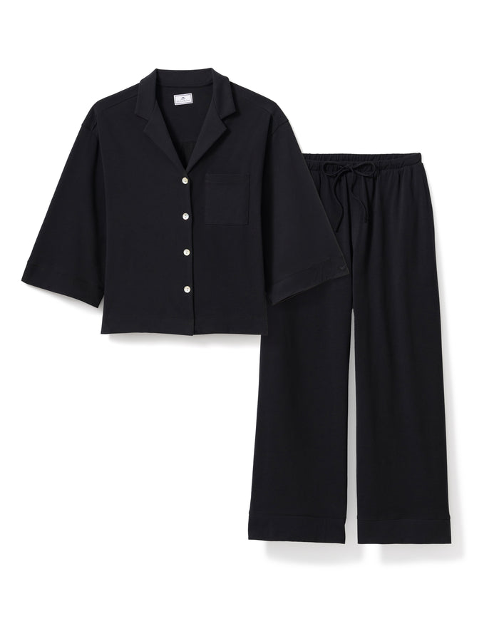 Modeling relaxed elegance, a person poses barefoot against an off-white background in The Olivia Wide Leg Pima Pajama Set from Petite Plume, featuring a loose-fitting black button-up top and wide-legged pants crafted from luxurious Peruvian Pima cotton.