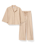 Against a plain backdrop, an individual with long curls and a relaxed stance wears The Olivia Wide Leg Pima Pajama Set in Camel by Petite Plume. The luxurious set features a button-up top and loose-fit pants, crafted from soft Pima cotton. They are barefoot, embodying comfort and elegance.