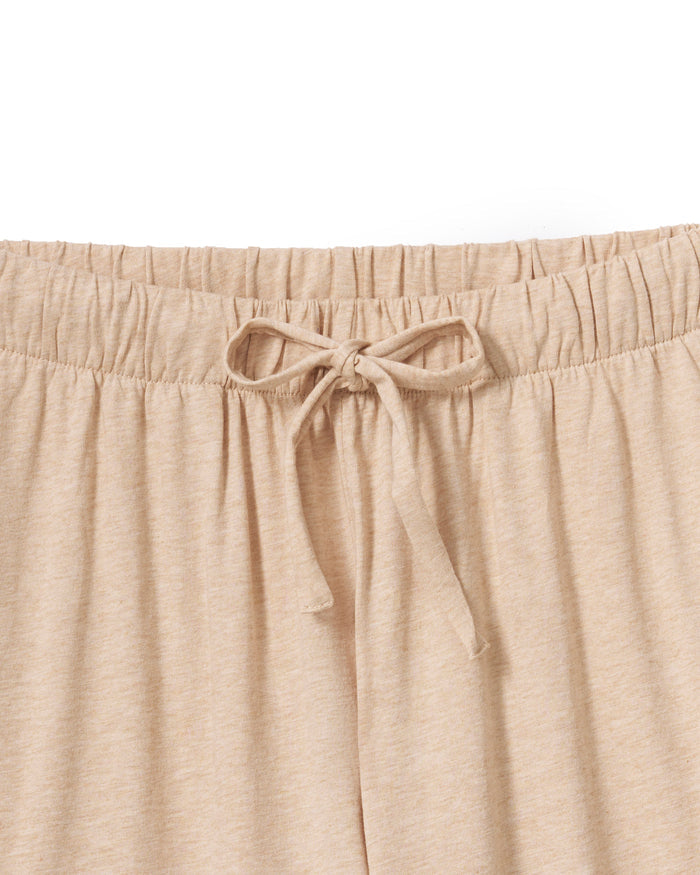 Against a plain backdrop, an individual with long curls and a relaxed stance wears The Olivia Wide Leg Pima Pajama Set in Camel by Petite Plume. The luxurious set features a button-up top and loose-fit pants, crafted from soft Pima cotton. They are barefoot, embodying comfort and elegance.