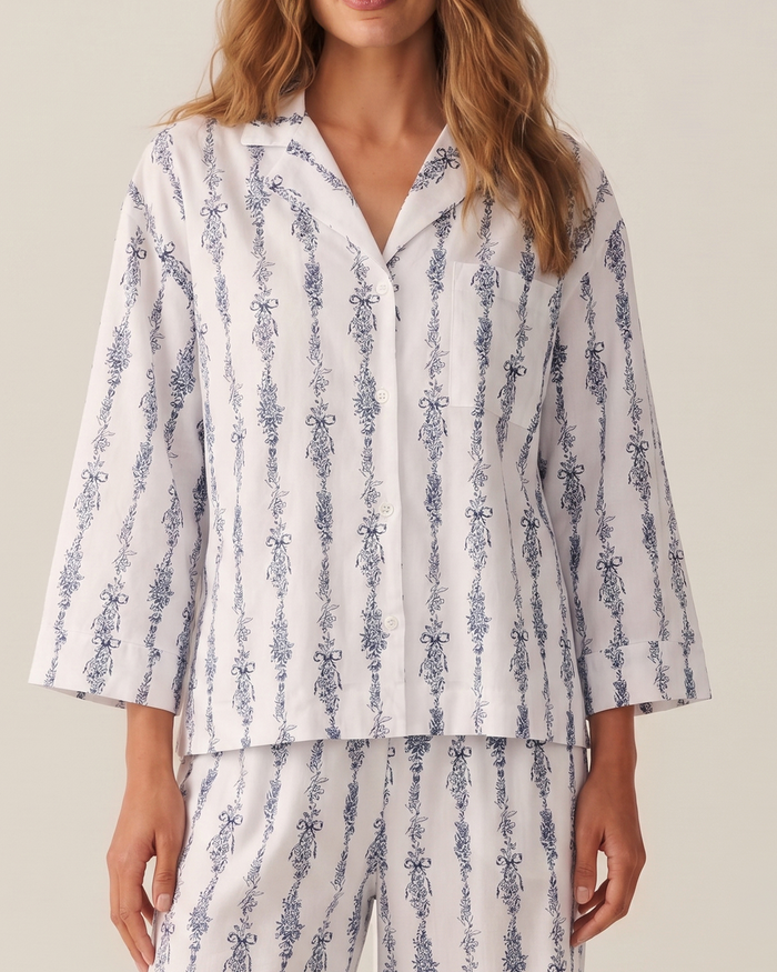 A woman wearing The Olivia Wide Leg Pima Pajama Set in Navy Mayfair Toile, which features a blue and white floral vertical stripe pattern. The pajama top has a collar, buttons, and long sleeves. She is shown from the shoulders to mid-thigh.