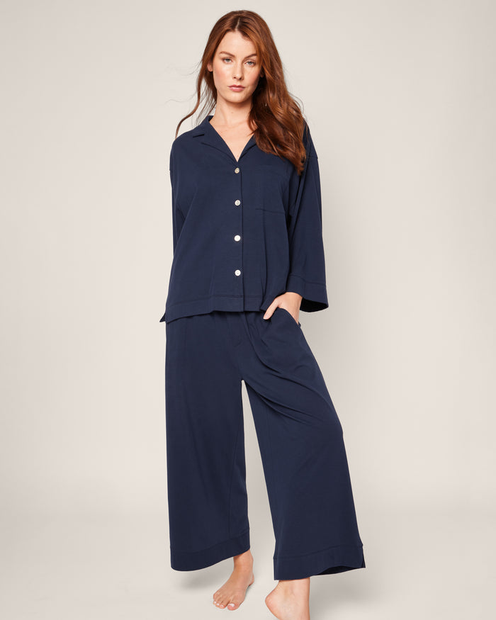 A person with long brown hair wears The Olivia Wide Leg Pima Pajama Set in Navy by Petite Plume, featuring a luxurious, buttoned, long-sleeved top and elegant wide-legged pants. They stand barefoot against an off-white background.