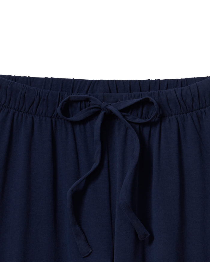 A person with long brown hair wears The Olivia Wide Leg Pima Pajama Set in Navy by Petite Plume, featuring a luxurious, buttoned, long-sleeved top and elegant wide-legged pants. They stand barefoot against an off-white background.