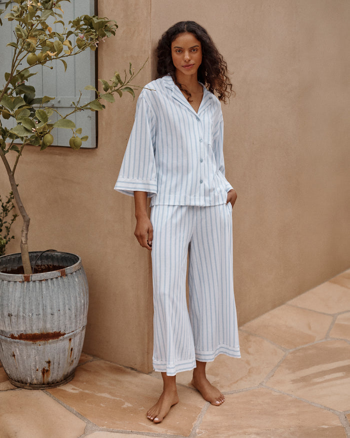 A person models the Petite Plumes Olivia Wide Leg Pima Pajama Set in Periwinkle Stripe, featuring a soft, luxurious white and light blue striped button-up long-sleeve shirt with matching wide-leg pants against a plain background that enhances its elegant simplicity.