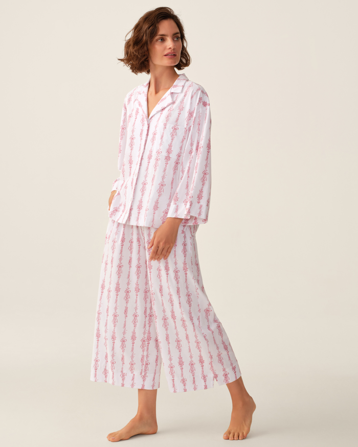 A person stands barefoot against a plain light background, wearing The Olivia Wide Leg Pima Pajama Set in Pink Mayfair Toile—loose-fitting, long-sleeved white pajamas adorned with pink vertical floral stripes.
