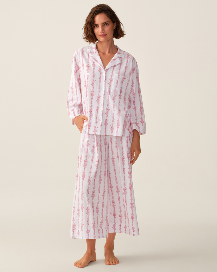 A woman stands barefoot against a plain background wearing The Olivia Wide Leg Pima Pajama Set in Pink Mayfair Toile—a loose-fitting, long-sleeve, button-up pajama top and matching wide-leg pants featuring a pink floral stripe pattern.