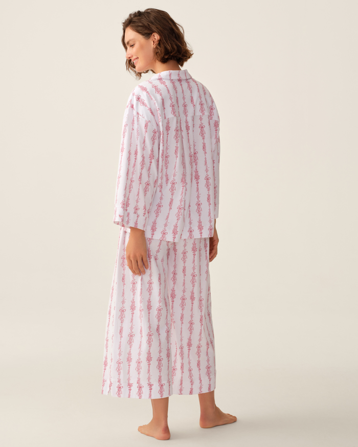 A woman stands barefoot, facing away, wearing The Olivia Wide Leg Pima Pajama Set in Pink Mayfair Toile. This set features loose, wide-leg pants and a long-sleeve top with a pink floral stripe pattern. The background is plain and light.