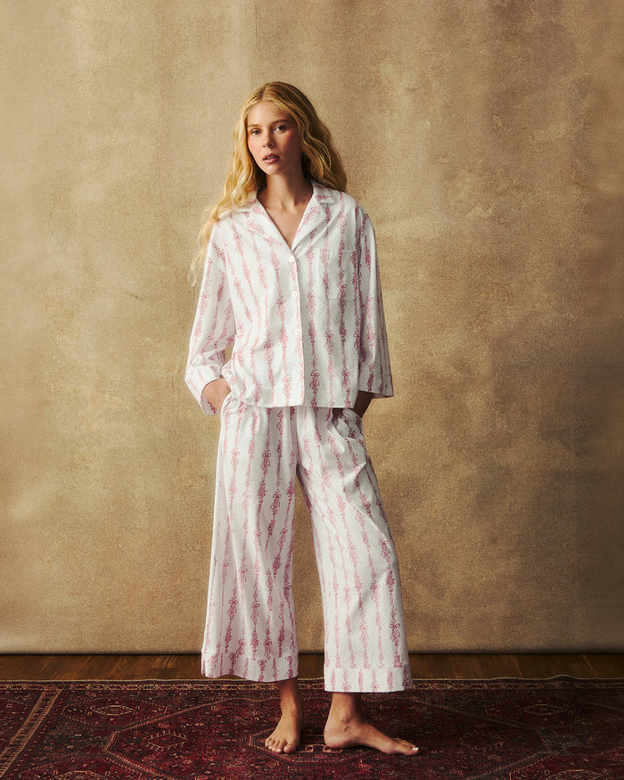 A person with long blonde hair stands barefoot on a red patterned rug, wearing The Olivia Wide Leg Pima Pajama Set in Pink Mayfair Toile, against a neutral, textured background.