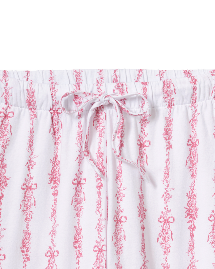 The Olivia Wide Leg Pima Pajama Set in Pink Mayfair Toile features wide-leg white pajama pants adorned with pink floral and bow vertical stripes, an elastic waistband, and a matching pink drawstring tied in a bow at the front.