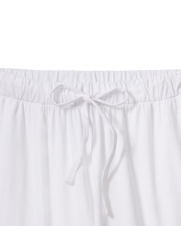 A person wears The Olivia Wide Leg Pima Pajama Set in White by Petite Plume, showcasing a luxurious Peruvian Pima cotton ensemble with a loose-fitting long-sleeve buttoned shirt and wide-leg pants. They hold a branch with green leaves, standing barefoot against a light background.