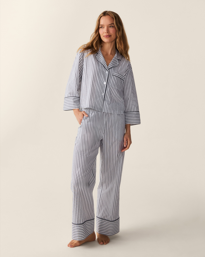 A woman stands barefoot, posing against a plain, light-colored background while wearing The Celine Poplin Pajama Set in Navy French Ticking, featuring blue and white stripes with long sleeves and pants.