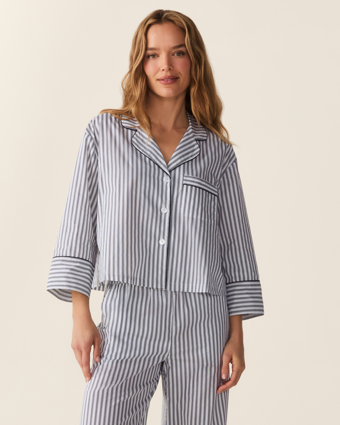 A woman wearing The Celine Poplin Pajama Set in Navy French Ticking, featuring blue and white vertical stripes, long sleeves, and a front pocket, stands smiling against a plain background.