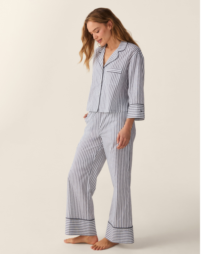 A woman stands barefoot against a plain light background, looking down and smiling while wearing The Celine Poplin Pajama Set in Navy French Ticking—featuring blue and white vertical stripes with long sleeves and pants.