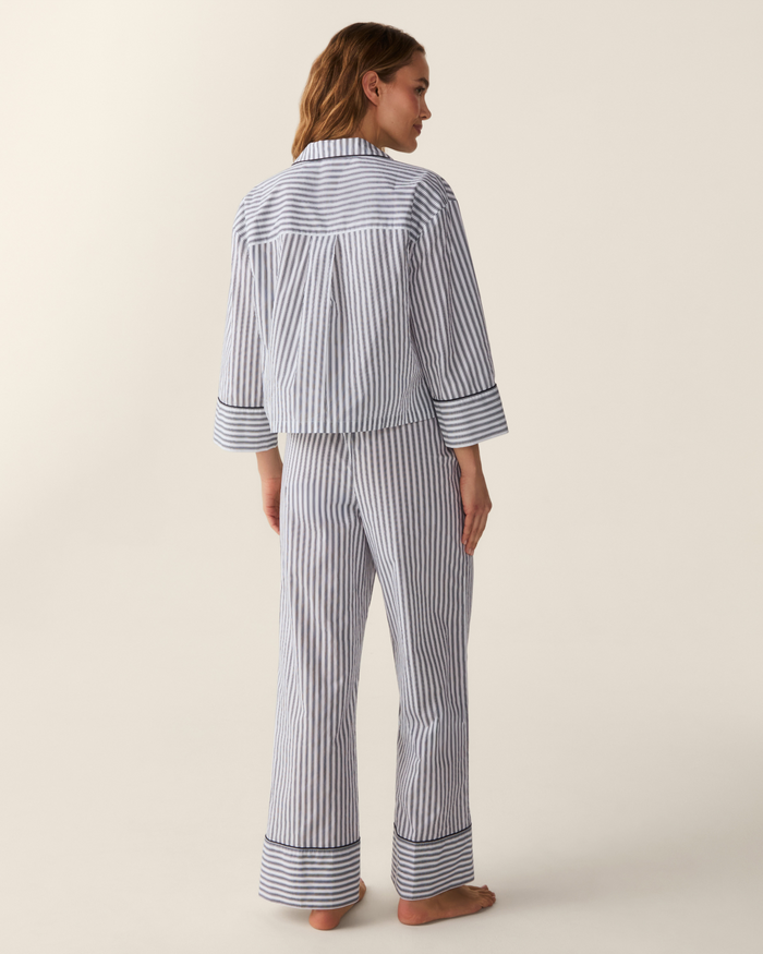 A woman stands barefoot, facing away, wearing The Celine Poplin Pajama Set in Navy French Ticking—featuring blue-and-white stripes with long sleeves and pants. The background is plain and light colored.